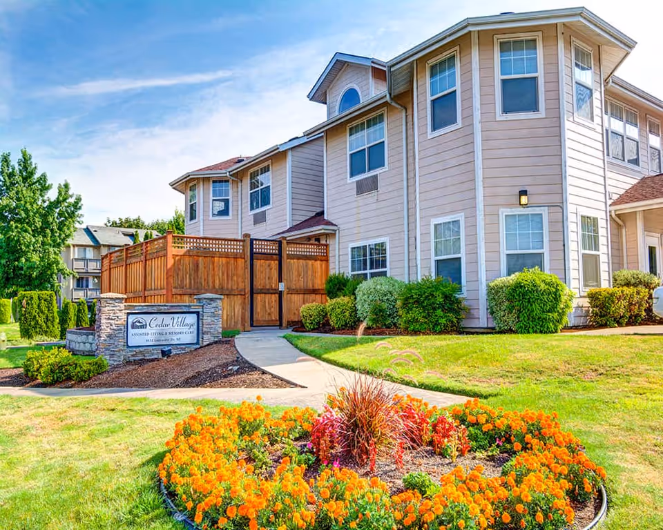Exterior view of Cedar Village by Cogir, an assisted living and memory care facility, showing a two-story beige building with multiple windows, a wooden fence, a curved sidewalk, and a landscaped garden with orange flowers and green shrubs under a blue sky.