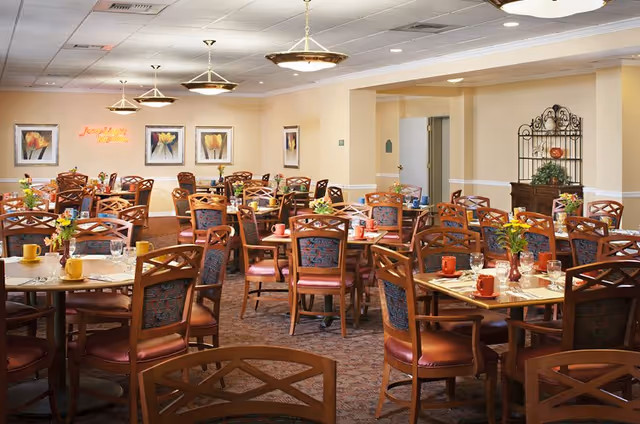 A spacious dining room in a senior living facility with multiple round wooden tables and chairs arranged neatly. Each table is set with colorful cups, glasses, and small flower arrangements. The walls are decorated with framed floral artwork and a neon sign that reads 'Simply Delicious'. The room has warm lighting from ceiling fixtures and a carpeted floor.