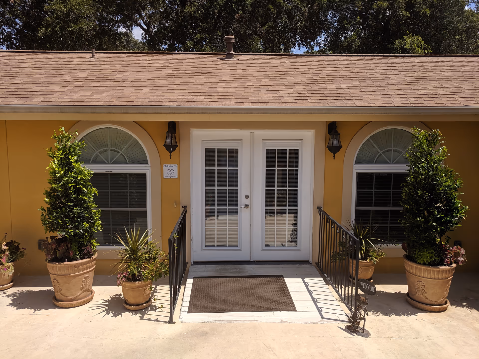 Front entrance of a building with double white doors and two arched windows on either side. There are potted plants and small trees flanking the entrance, with a welcome sign on the right side. The building exterior is painted yellow with a brown shingled roof.