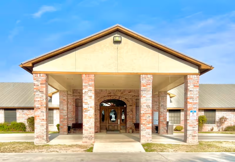Front exterior view of Amistad Nursing & Rehabilitation Center showing a covered entrance supported by brick columns, with double wooden doors and windows on either side under a clear blue sky.