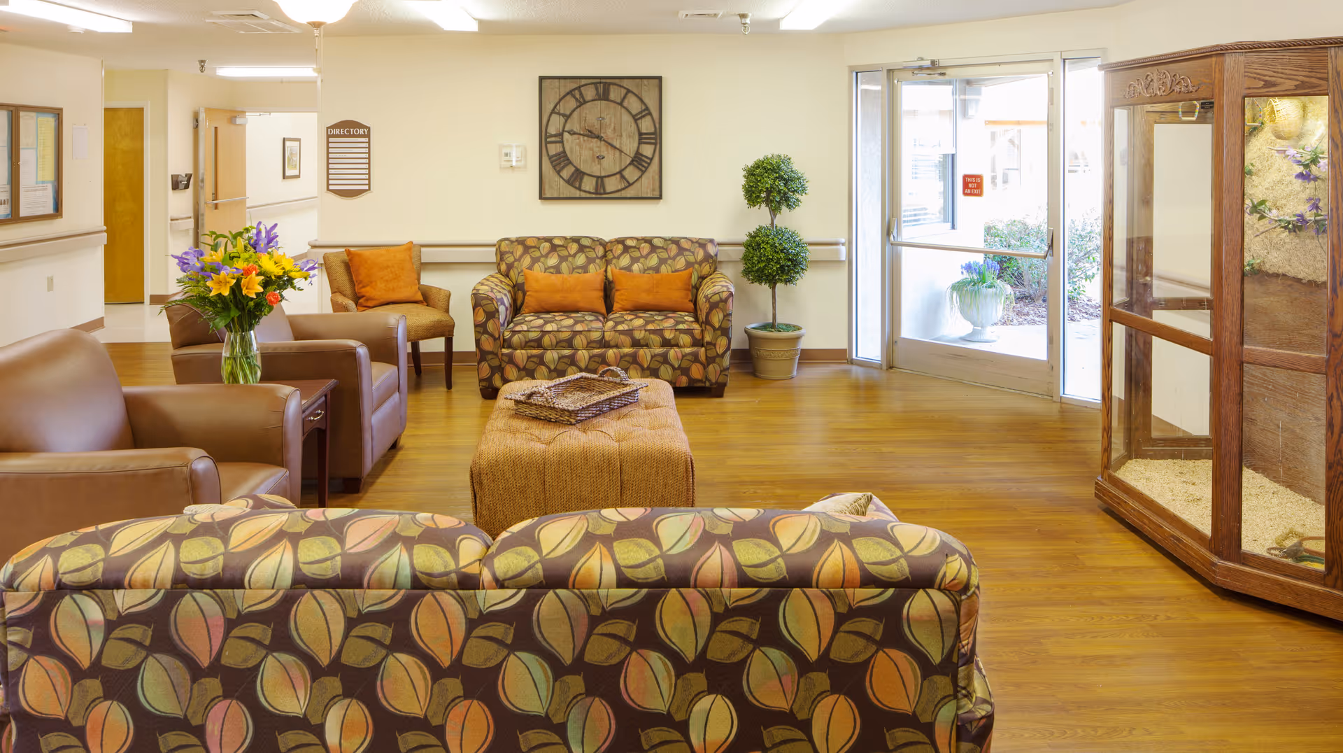 A bright and welcoming senior living facility common area with patterned sofas, leather armchairs, a large ottoman, a vase of colorful flowers on a side table, a large wall clock, potted plants, and glass doors leading outside.
