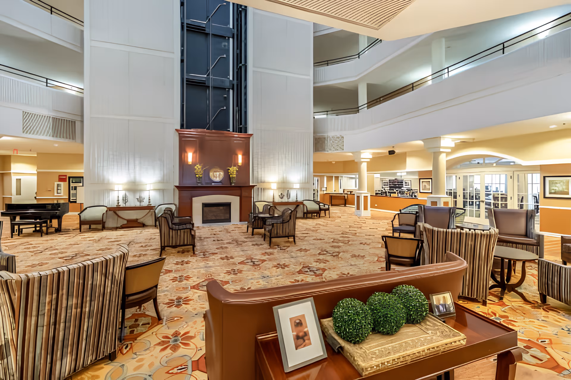 Spacious and well-lit senior living facility common area with multiple seating arrangements including armchairs and sofas, a fireplace with decorative plants and framed pictures on a wooden table in the foreground, and a grand piano on the left side. The area features high ceilings with balconies overlooking the space and warm lighting.