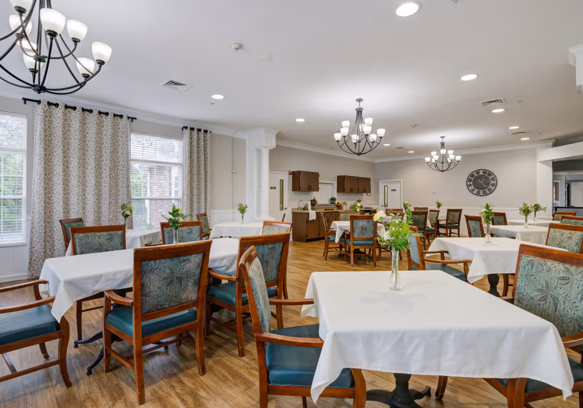 A bright dining room in a senior living community with multiple tables covered in white tablecloths, each adorned with a small vase of flowers. Wooden chairs with green upholstered seats and backs surround the tables. The room features large windows with patterned curtains, wooden flooring, and several black chandeliers with white lampshades hanging from the ceiling. A large wall clock is visible on the far wall.