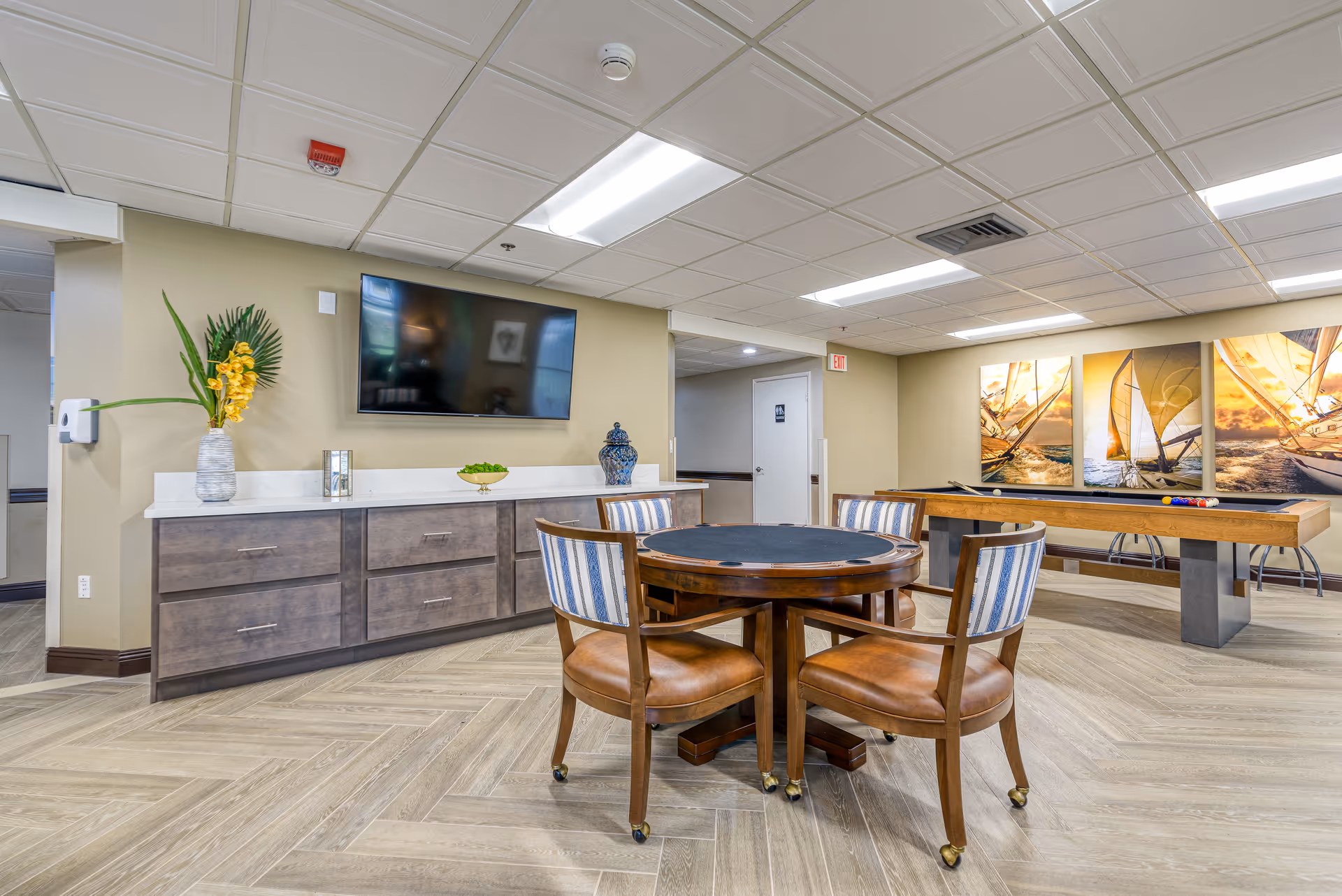Bright senior living common room with a round game table and chairs, wall-mounted TV, cabinets and a shuffleboard table.