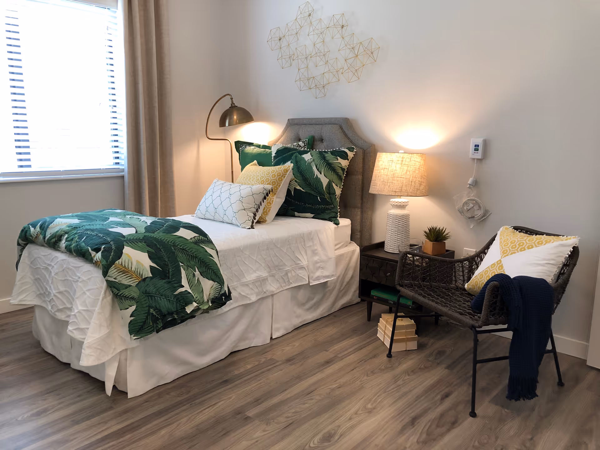 A cozy bedroom with a single bed adorned with white sheets and green leaf-patterned pillows and blanket. Next to the bed is a black nightstand with a white textured lamp and a small potted plant. A black woven chair with a yellow and white pillow and a dark blue throw blanket is placed beside the nightstand. The room has light-colored walls, a window with blinds and beige curtains, and wooden flooring.