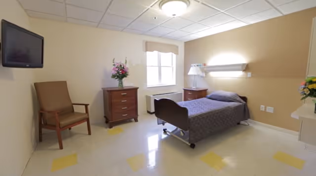 Furnished assisted living bedroom with a single hospital-style bed, nightstands, dresser, armchair, wall-mounted TV and a window.