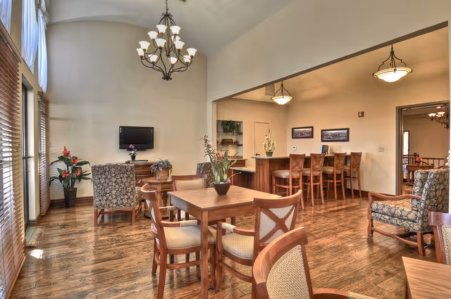 A spacious and well-lit common area with wooden flooring, featuring a square wooden table with four cushioned chairs in the foreground. In the background, there are two upholstered armchairs facing a wall-mounted flat-screen TV. To the right, a counter with several high wooden stools is visible under hanging light fixtures. Large windows with blinds allow natural light to fill the room, and decorative plants and framed pictures add to the cozy ambiance.