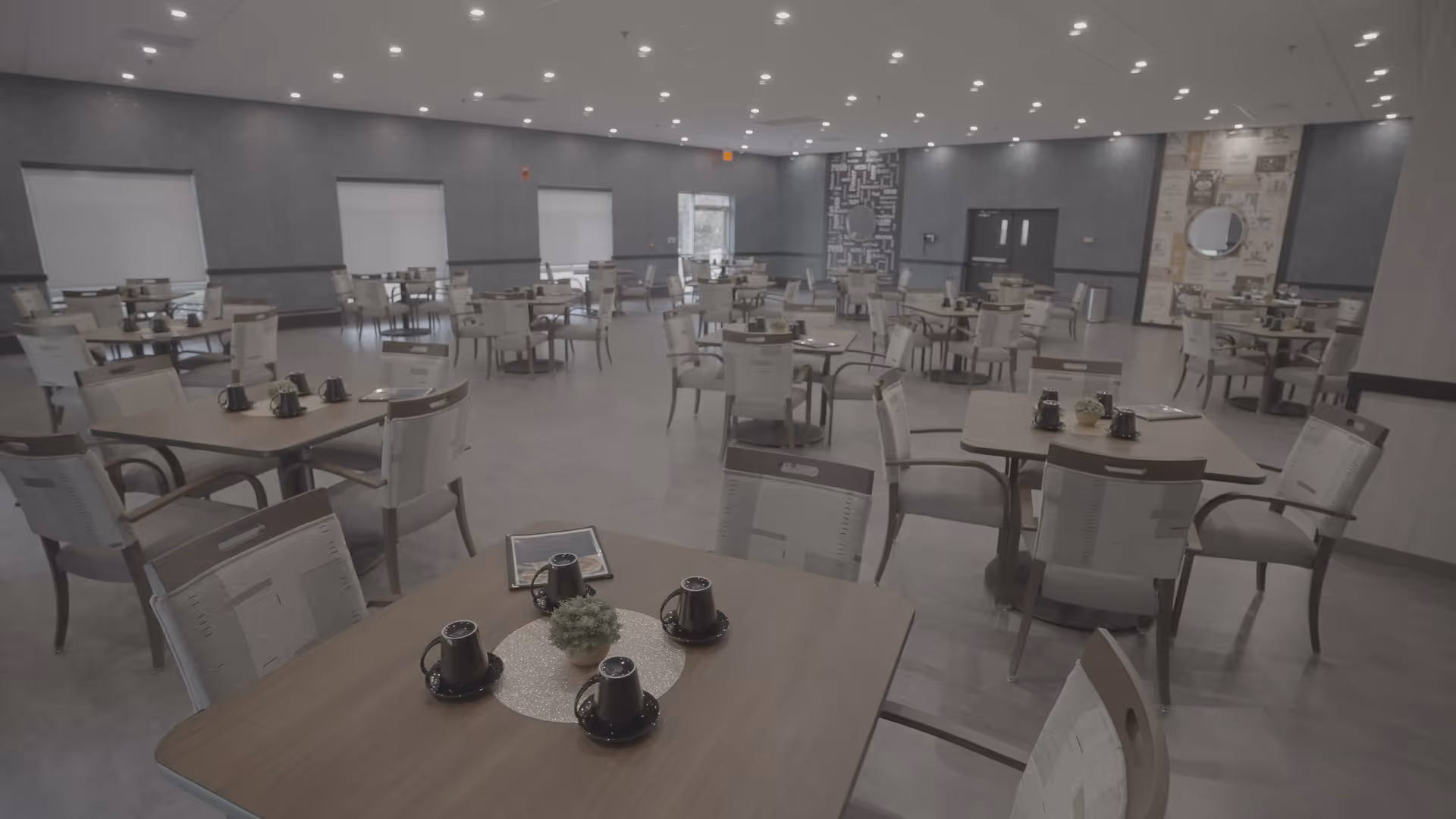 A spacious dining room with multiple square tables, each surrounded by four chairs. The tables are set with black cups and small decorative plants. The room has large windows with blinds, neutral-colored walls, and a patterned accent wall with a round mirror. The floor is light wood, and the ceiling has recessed lighting.