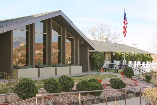 Exterior view of Hurricane Health And Rehabilitation facility showing a building with large triangular windows, a landscaped garden with bushes and rocks, a white picket fence, a bench, and an American flag on a flagpole.
