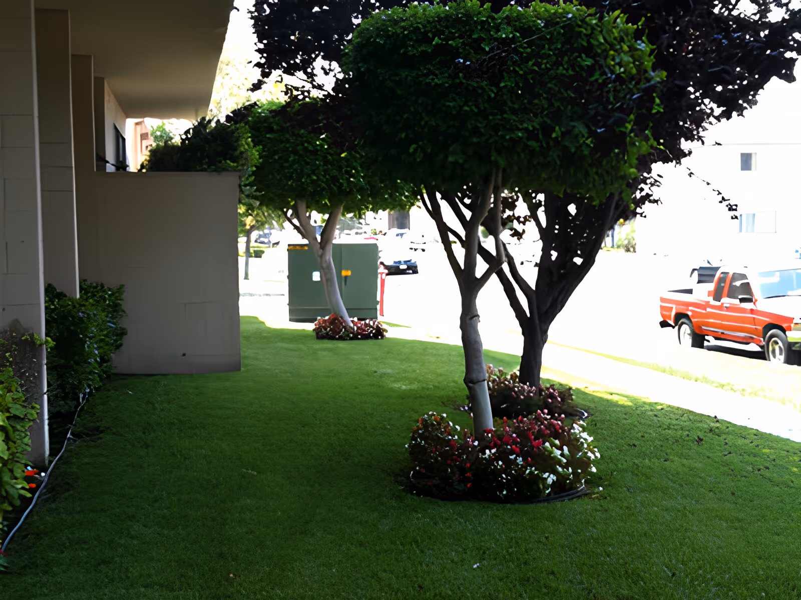 Outdoor view of a grassy area with neatly trimmed trees and flower beds next to a building. A red pickup truck is parked on the street in the background.