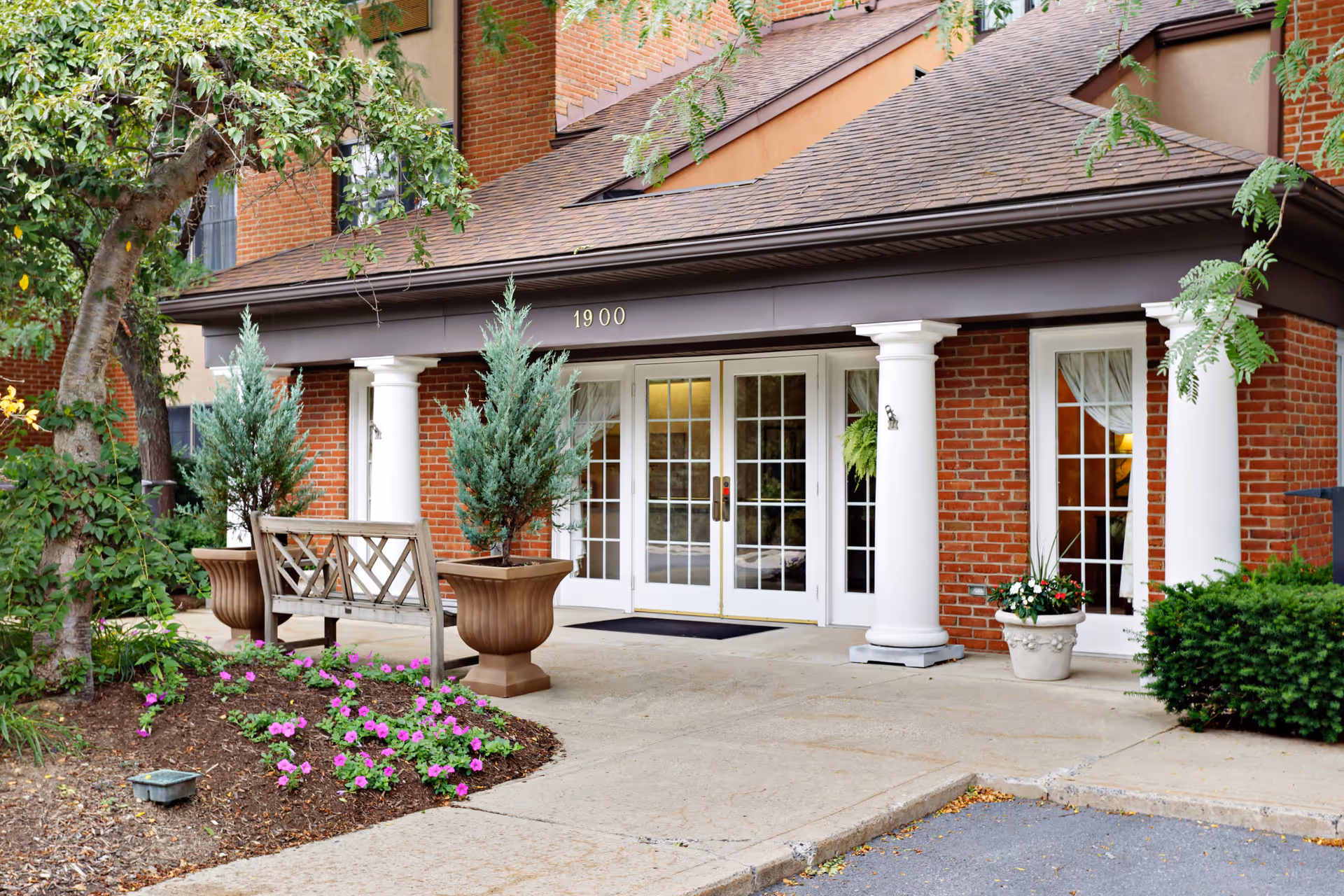 Entrance of a senior living facility with a brick exterior, white columns, glass double doors, potted plants, a wooden bench, and a small garden with flowers.