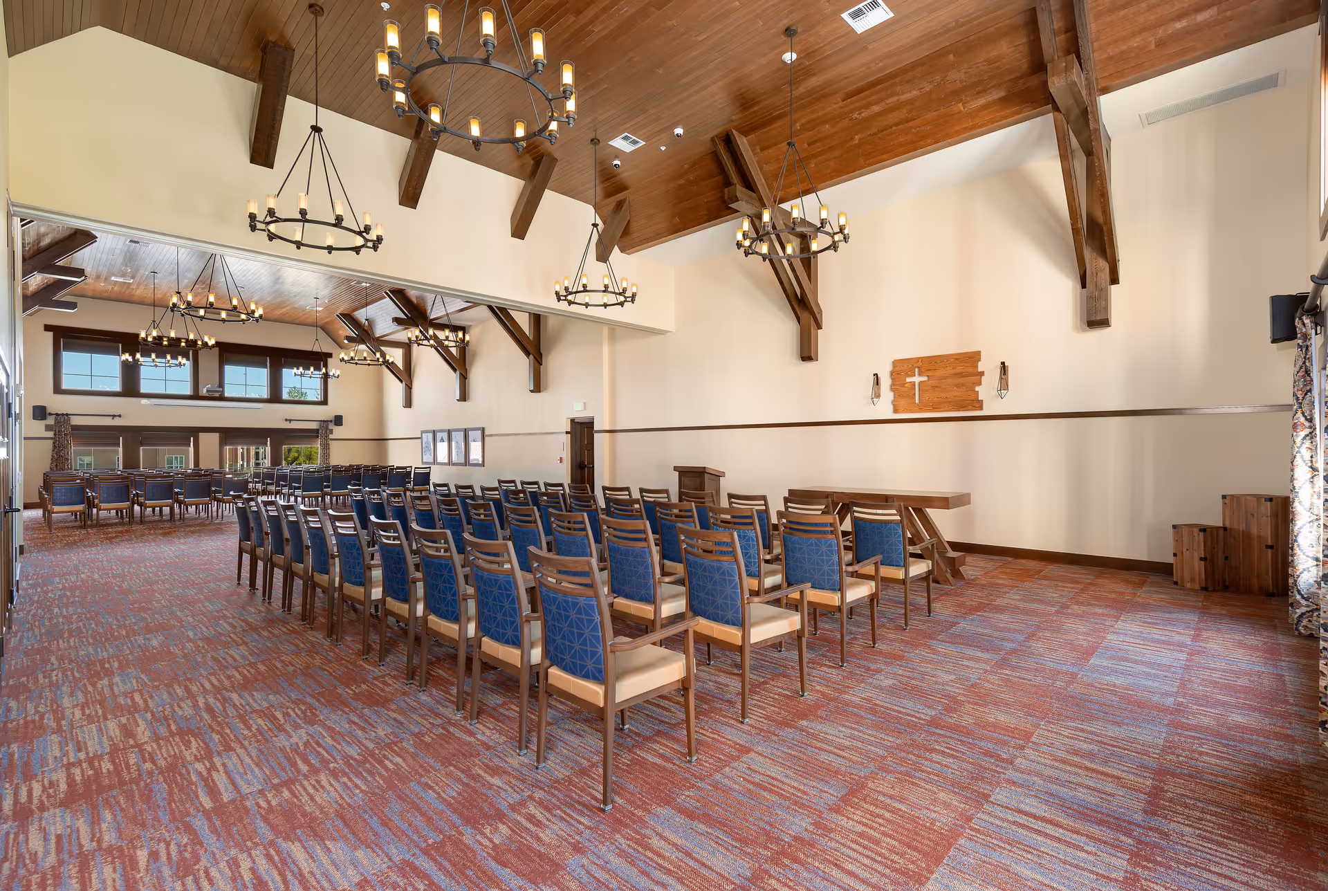 A spacious room with rows of wooden chairs with blue cushions arranged facing a wooden podium and table. The room has high vaulted wooden ceilings with exposed beams and multiple circular chandeliers. The walls are light-colored with a wooden cross mounted on one wall. Large windows and patterned curtains are visible on the right side, and the carpet has a multicolored pattern in shades of red and blue.