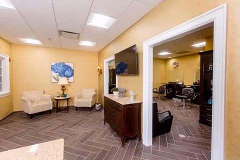 Interior view of a senior living facility room with beige walls and brown herringbone tile flooring. The room features two patterned armchairs with a small round table and lamp between them, a wall-mounted flat screen TV above a wooden cabinet, and an open doorway leading to another room with salon chairs and mirrors.