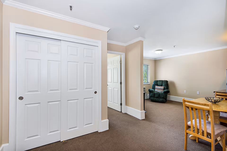 Interior view of a senior living facility room with beige walls and carpeted floor. The room features a white double-door closet on the left, an open doorway leading to another room, a green upholstered armchair near a window with blinds, and a wooden dining table with chairs on the right.