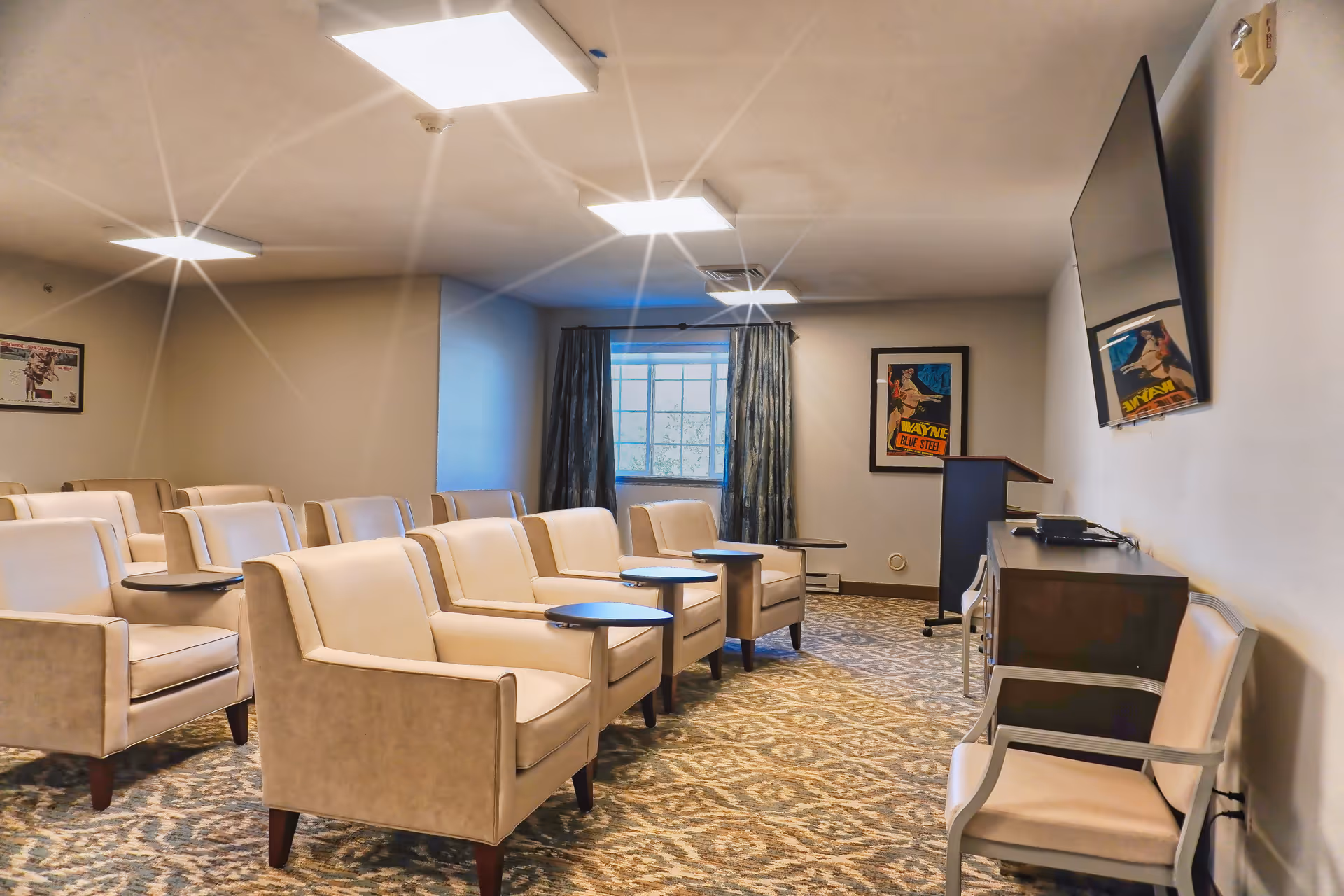 A small theater-style room with several beige armchairs arranged in rows, each with a small attached table. The room has patterned carpet, a large window with curtains, framed posters on the walls, a podium, and a large flat-screen TV mounted on the wall.