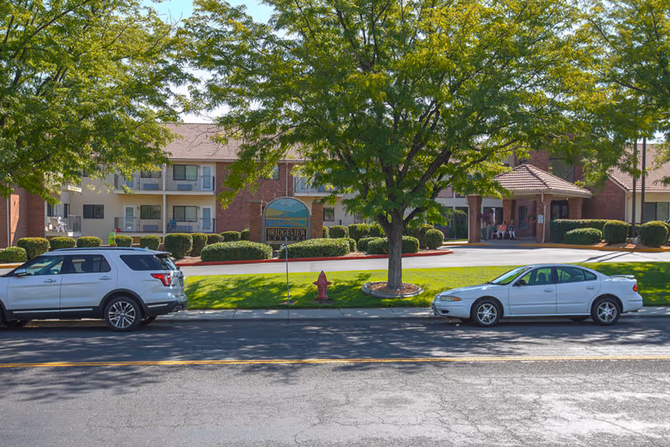Exterior view of Bridgeview Estates senior living facility with two parked cars in front, a large tree in the center, and a brick building with balconies and a covered entrance in the background.