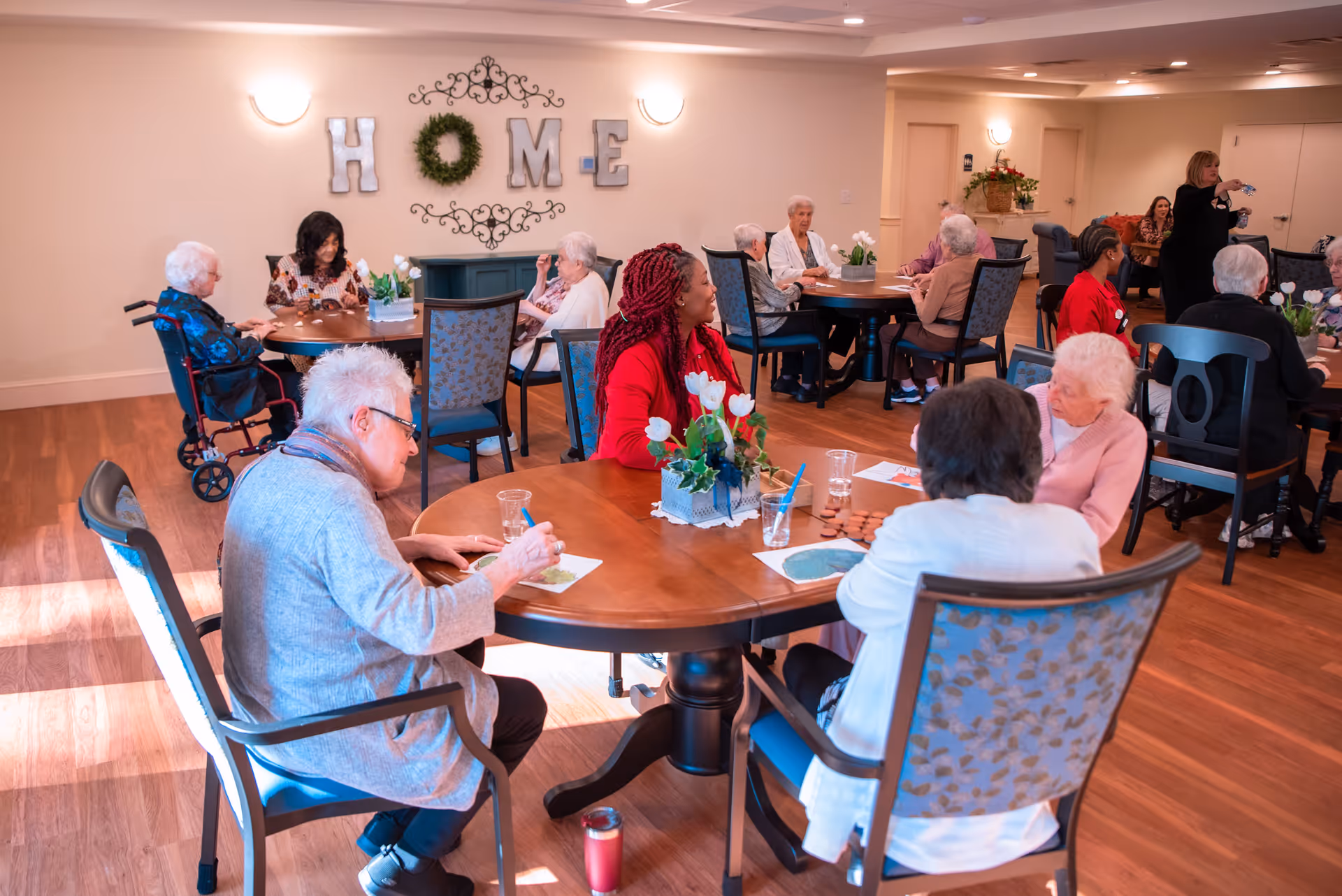 A group of elderly people and caregivers sitting around multiple round tables in a well-lit common room with wooden floors. The walls are decorated with the word 'HOME' and a wreath. Some people are engaged in activities, while others are conversing.
