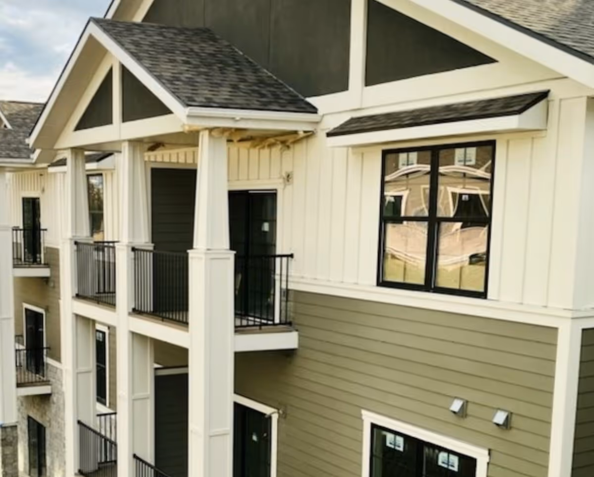 Exterior view of a multi-story residential building with balconies, white columns, and a combination of white and olive green siding under a gabled roof.