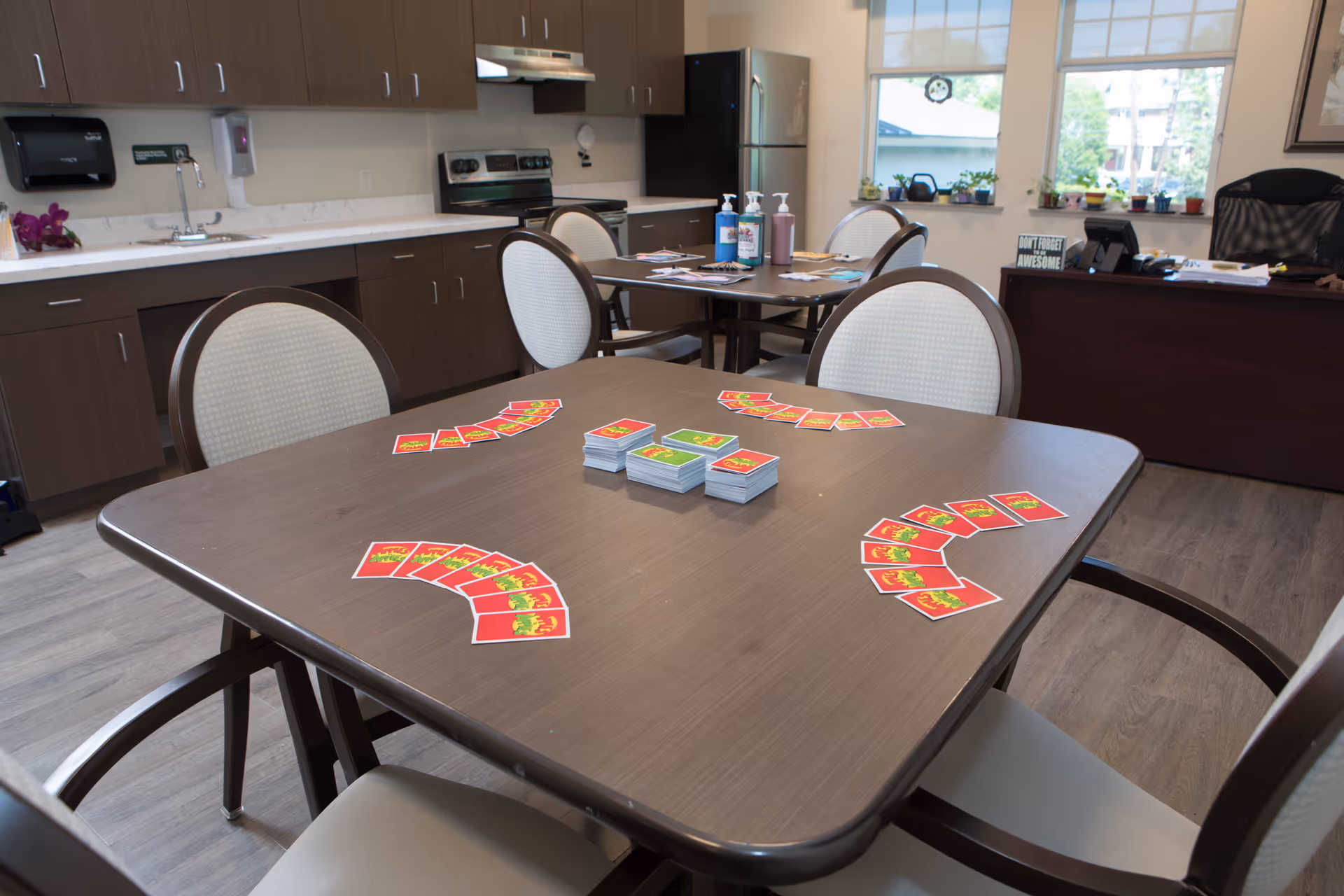 A room with a wooden table set for a card game with four sets of colorful cards arranged in front of four chairs. In the background, there is a kitchenette with cabinets, a stove, a refrigerator, and a sink. Another table with hand sanitizer bottles and papers is visible, along with a desk and office chair near windows with blinds.
