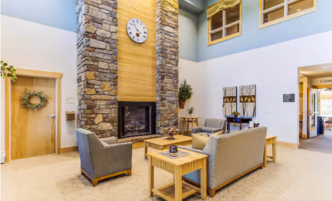 A cozy senior living facility common area with a tall stone fireplace featuring a clock above it. The room has comfortable seating including two armchairs and a sofa arranged around a wooden coffee table and side tables. The walls are light-colored with some decorative artwork and plants. There are windows near the ceiling and a door with a wreath on it to the left.