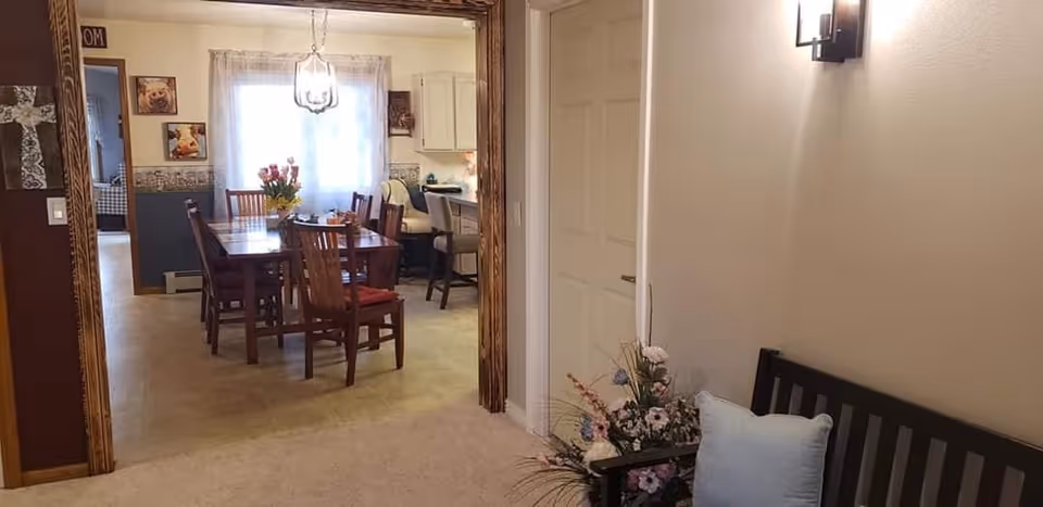 View of a dining room with a wooden table and chairs, a vase with flowers on the table, and a window with sheer curtains letting in natural light. Adjacent to the dining room is a kitchen area with bar stools. In the foreground, there is a bench with a cushion and a floral arrangement next to it, along with a wall sconce providing light.