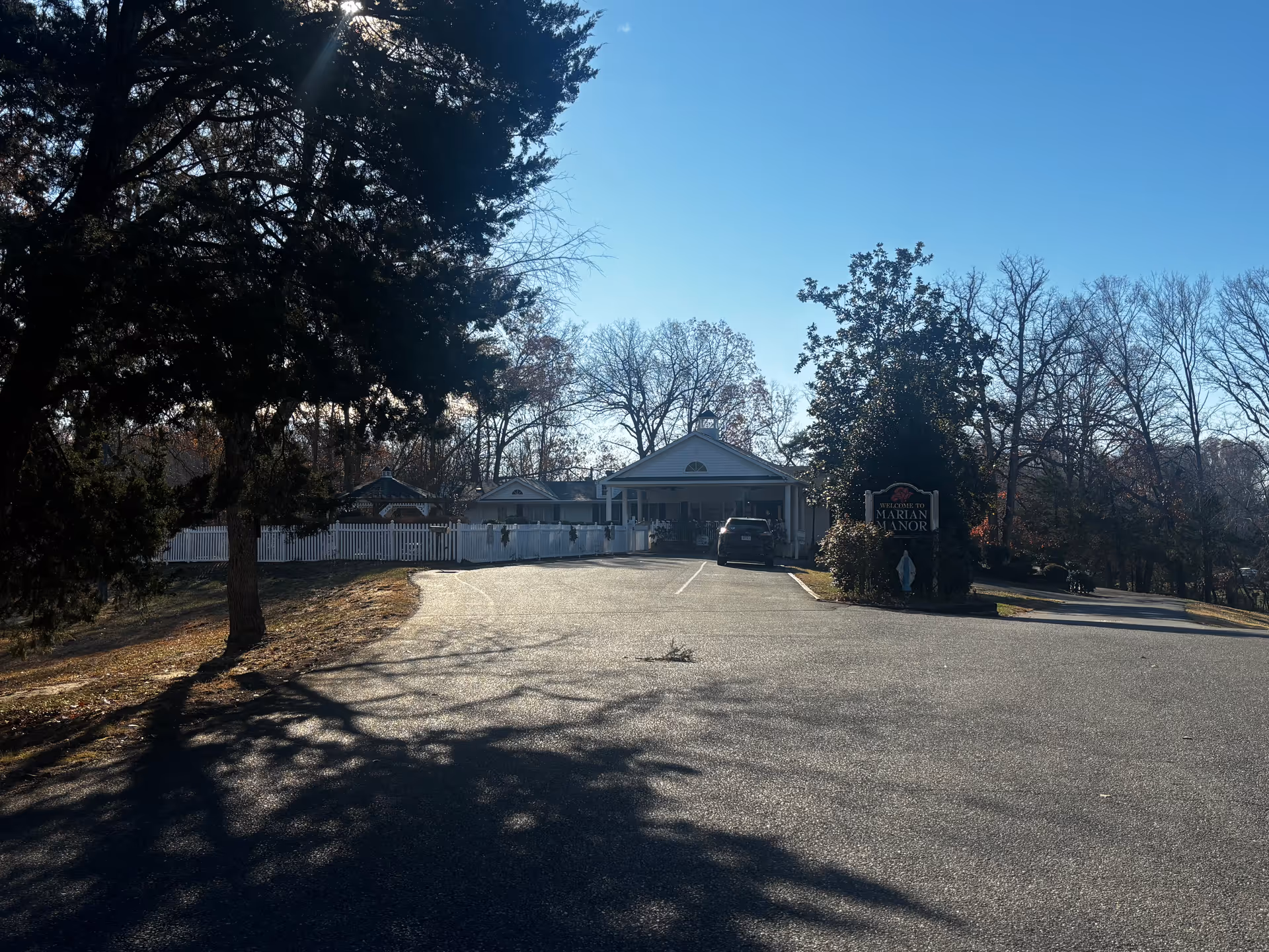 Driveway leading to the front entrance of Marian Manor, a single-story white building with a porch, parking area and surrounding trees.