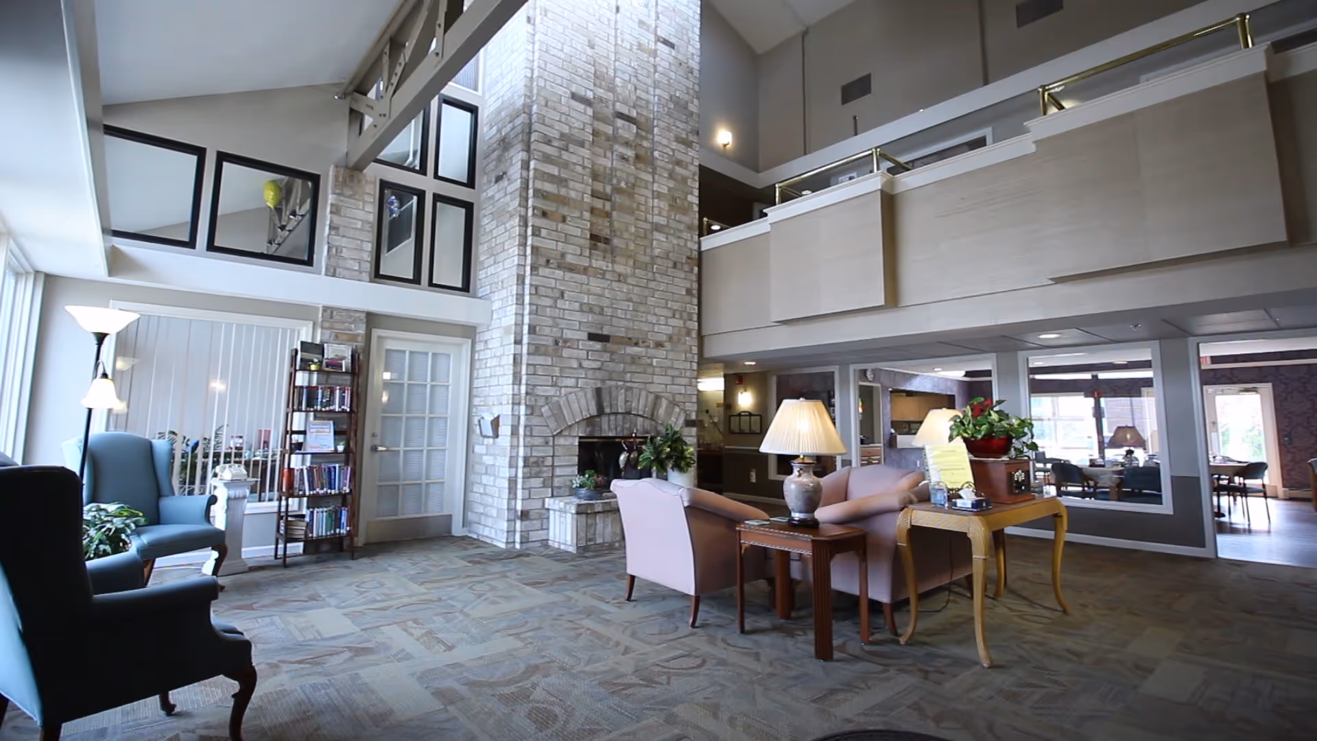 A spacious assisted living facility common area with high ceilings and a large brick fireplace in the center. The room features comfortable seating including armchairs and sofas, a wooden side table with a lamp, a small bookshelf, and large windows allowing natural light to fill the space. There is a second-floor balcony overlooking the area and a table with a plant and informational materials.