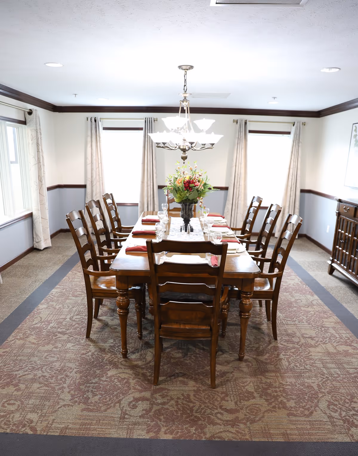 A formal dining room with a long wooden table set for eight people. The table has a floral centerpiece, glassware, and folded napkins. The room has three windows with light-colored curtains, a patterned carpet, and a chandelier hanging above the table.