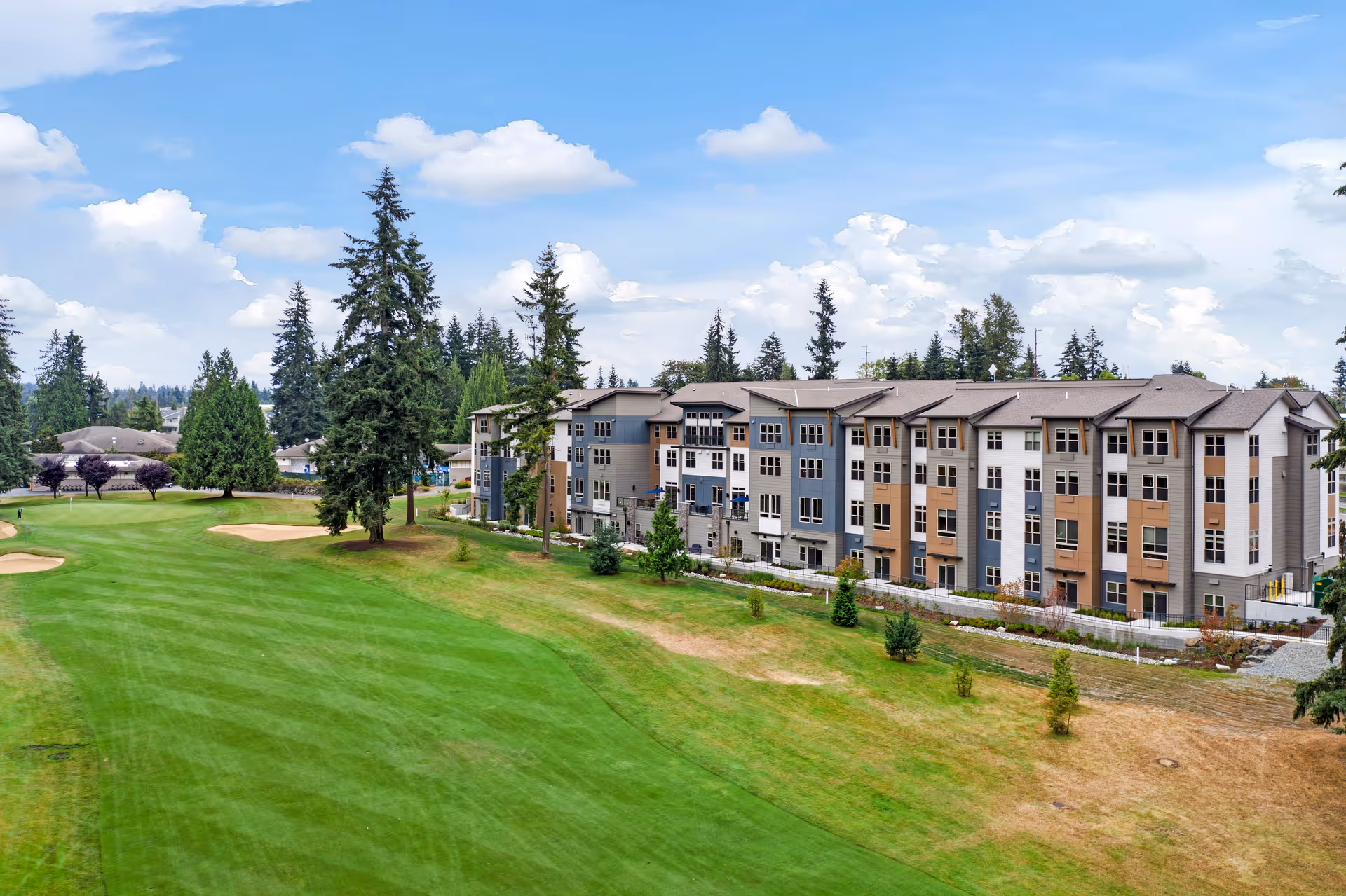 Exterior view of a multi-story senior living facility building with a well-maintained green lawn and tall trees under a partly cloudy sky.
