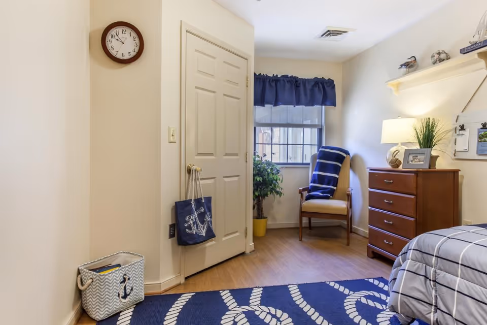 A cozy bedroom corner in a senior living facility featuring a wooden dresser with a lamp, framed photo, and plant on top. Next to the dresser is a cushioned chair with a blue and white striped blanket draped over it, positioned near a window with a navy blue valance. A door with a blue tote bag hanging on the handle is visible, along with a wall clock above it. The floor has a blue rug with a white rope pattern, and a basket with an anchor design sits in the corner.