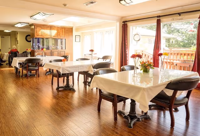 A bright dining room with wooden floors and several tables covered with white tablecloths. Each table has a small vase with flowers. Chairs are arranged around the tables, and large windows with red curtains allow natural light to fill the room. In the background, a person in a wheelchair is visible near a wooden cabinet.