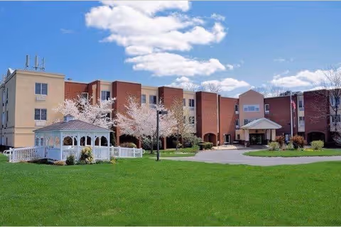 Front exterior of a multi-story senior living building with a white gazebo, green lawn, and blossoming trees under a blue sky.