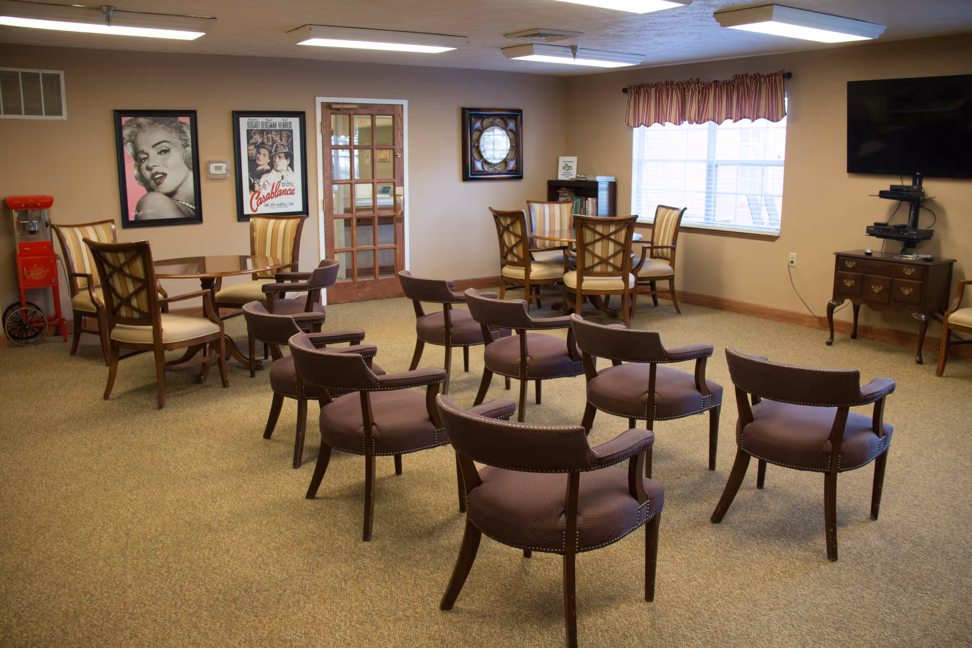 A common room with multiple chairs arranged in rows facing a television mounted on the wall. There are two round tables with chairs around them near the walls, a popcorn machine in the corner, framed movie posters on the wall, and a window with striped curtains letting in natural light.