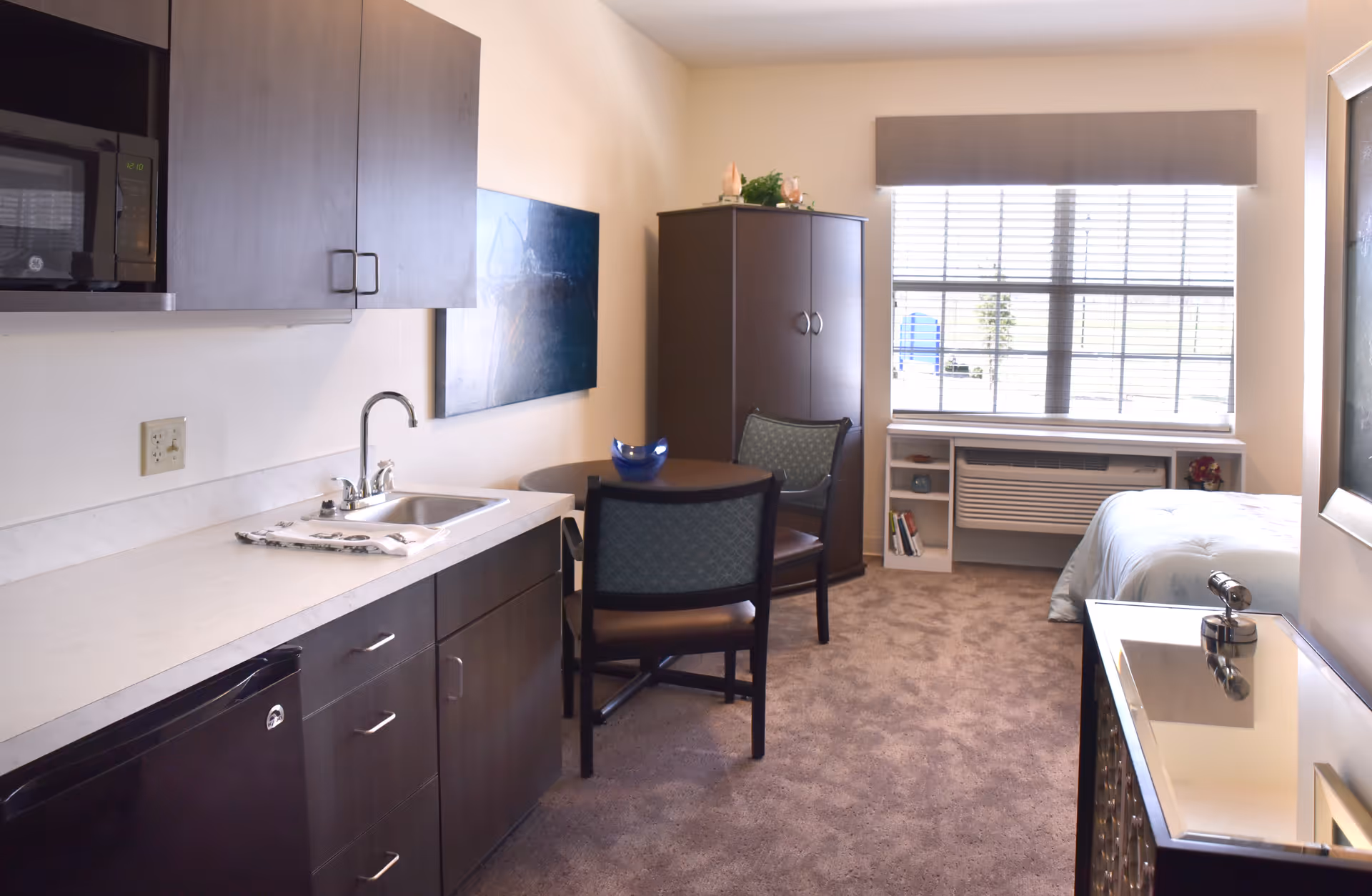 Studio-style senior living interior with a kitchenette, small round dining table, wardrobe and a bed by a large window.