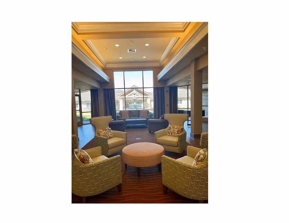 A cozy seating area in a senior living facility with four armchairs arranged around a round ottoman. The chairs have patterned upholstery and decorative pillows. In the background, there is a large window with a view of the building exterior and a sofa with additional seating. The room has warm lighting and a high ceiling with recessed lights.