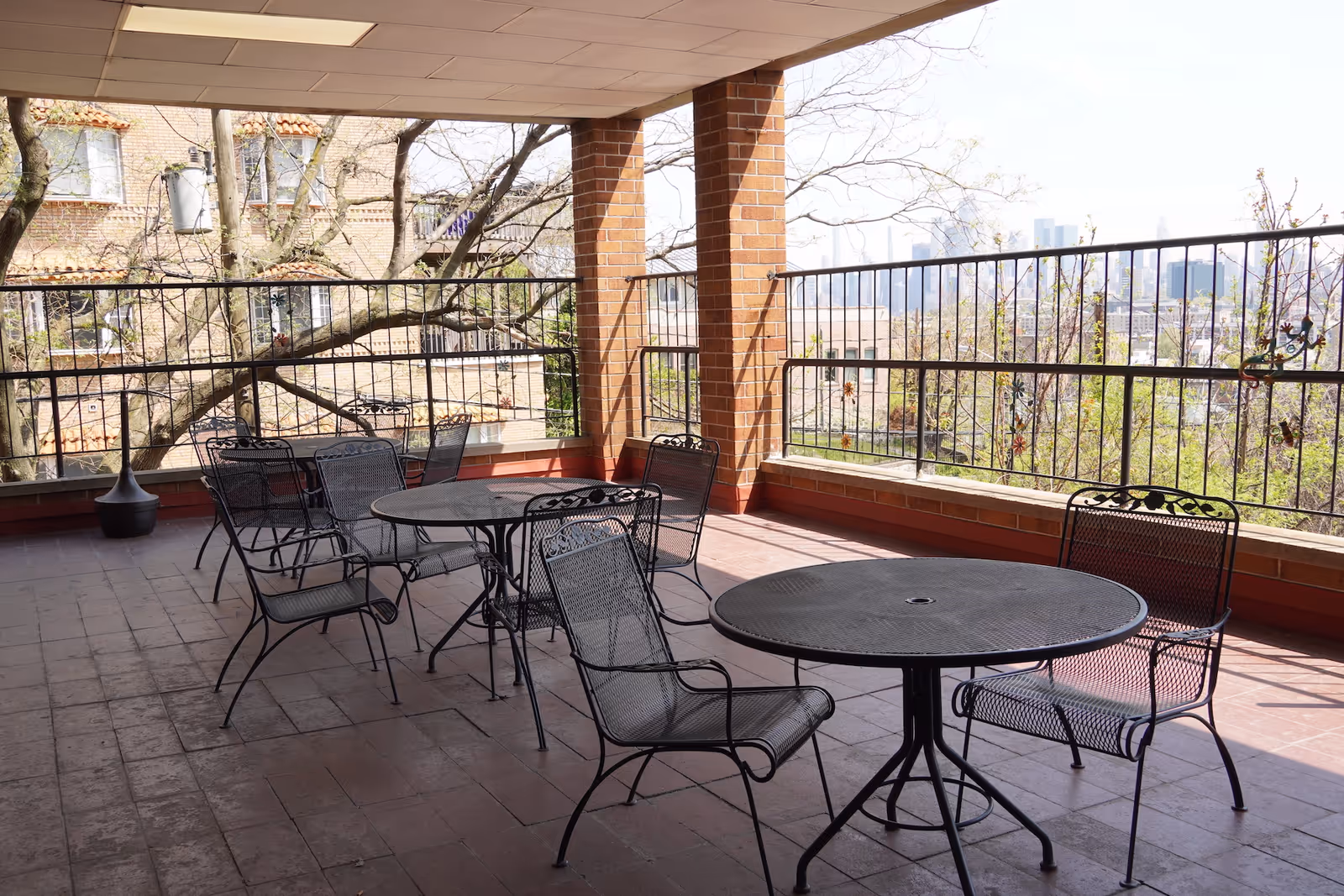 Covered outdoor patio area with several black metal tables and chairs arranged on a tiled floor. The patio is enclosed by a black metal railing decorated with small ornamental designs. Brick pillars support the roof, and trees and buildings are visible in the background.