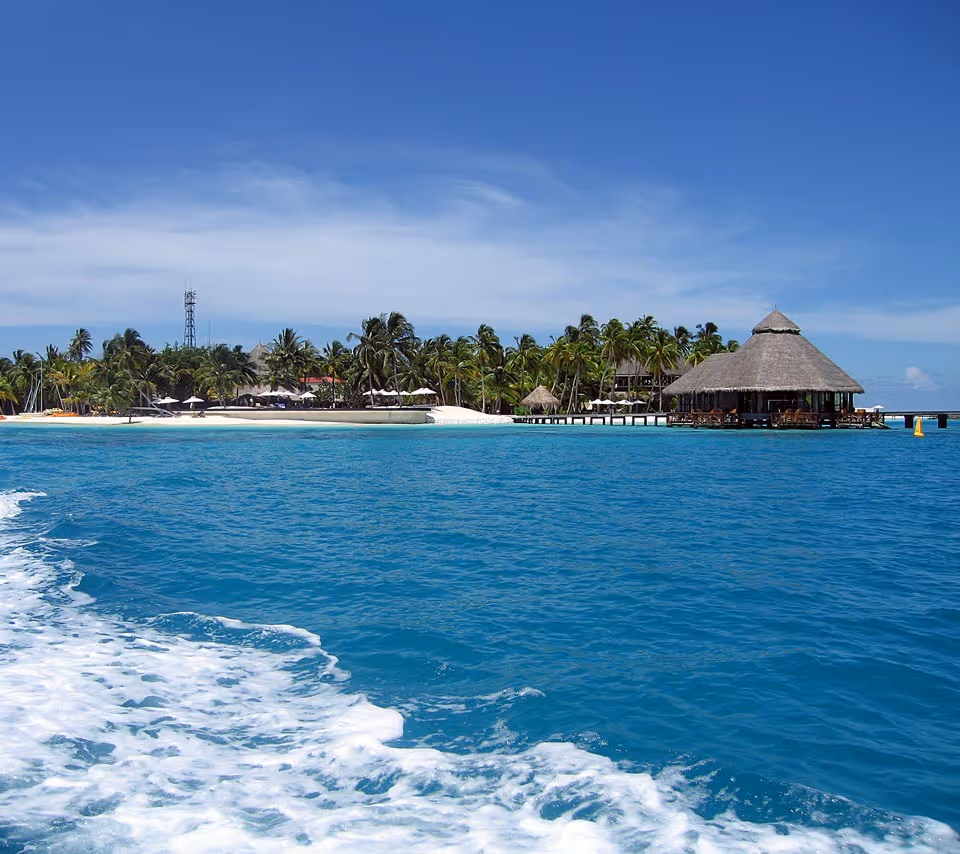 View of a tropical island with palm trees, a sandy beach, and a thatched-roof building on stilts over clear blue water under a partly cloudy sky.