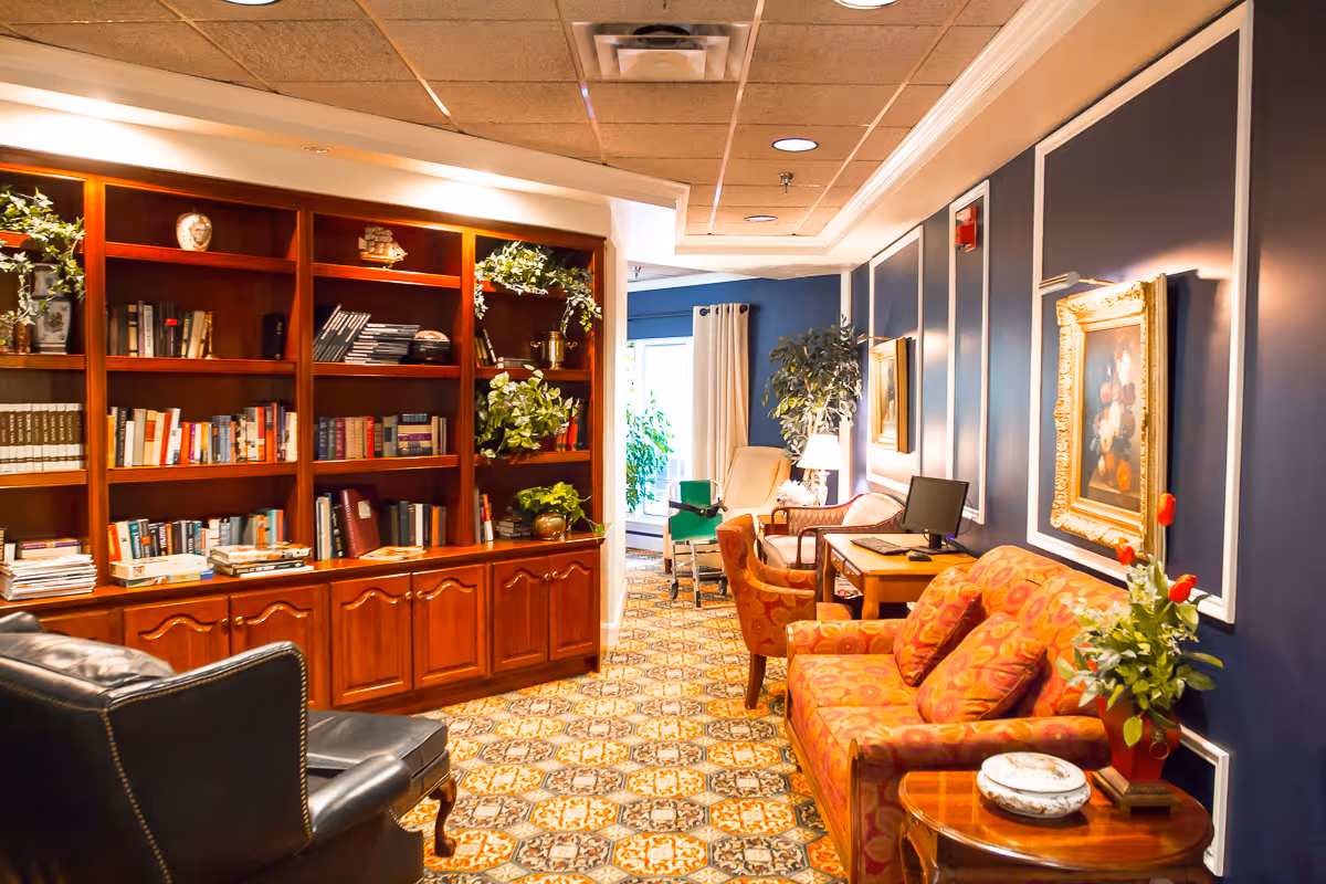 A cozy interior lounge area with a patterned carpet, a wooden bookshelf filled with books and decorative plants, a black leather armchair, a red patterned sofa with matching cushions, a wooden side table with a plant and decorative items, a desk with a computer, and framed artwork on dark blue walls.