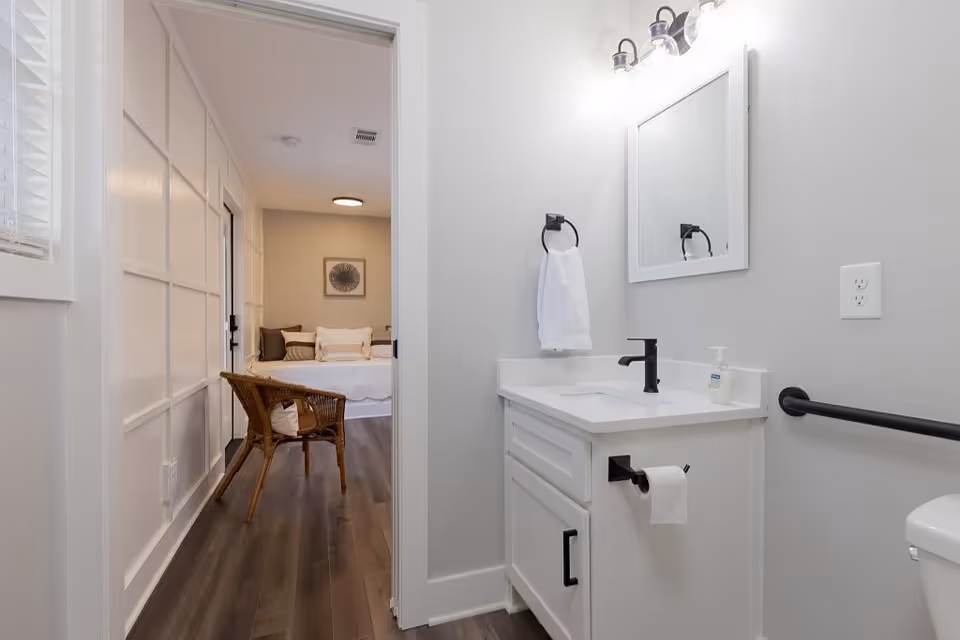 View of a small bathroom with a white vanity, black faucet, and a mirror above. A towel ring with a white hand towel and a toilet paper holder are mounted on the wall. The bathroom opens into a bedroom with a wicker chair and a bed with pillows, visible through the doorway.