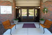 Brick entrance to Kimes Nursing & Rehab Center showing double glass doors, a maroon mat, two benches and potted plants.