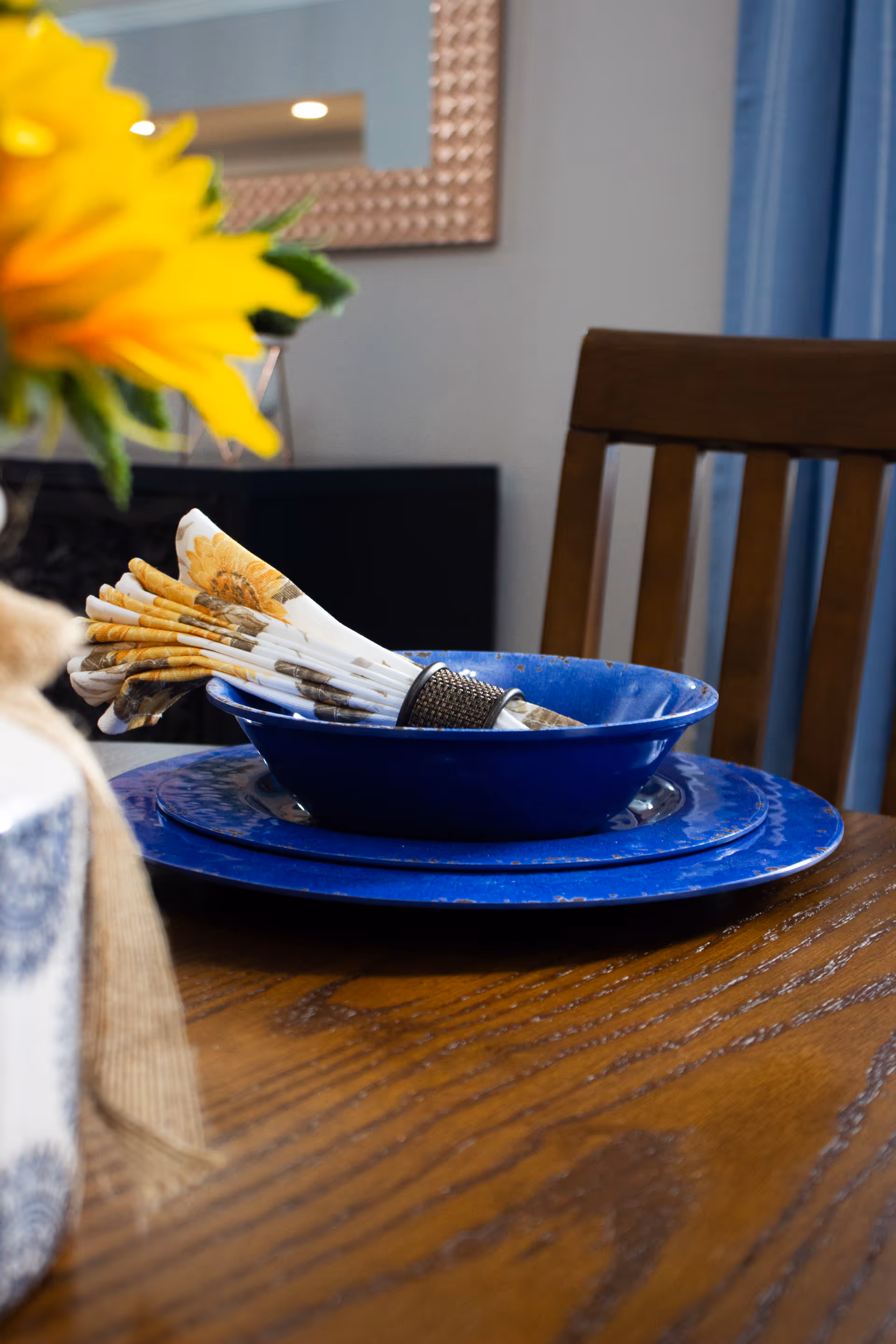 Close-up of a dining table setting with a blue plate and bowl, a folded sunflower-patterned napkin held by a napkin ring, a wooden chair in the background, and a blurred yellow flower in the foreground.