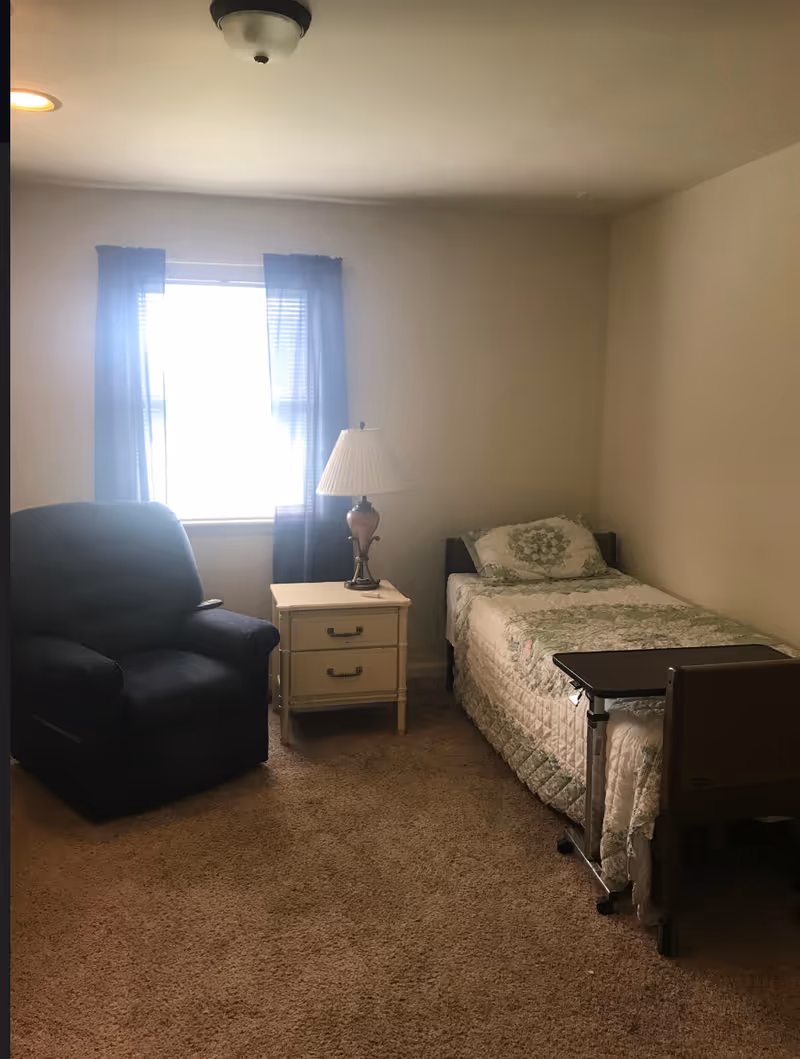 A small bedroom with a single bed, nightstand, lamp, recliner, and window with blue curtains.