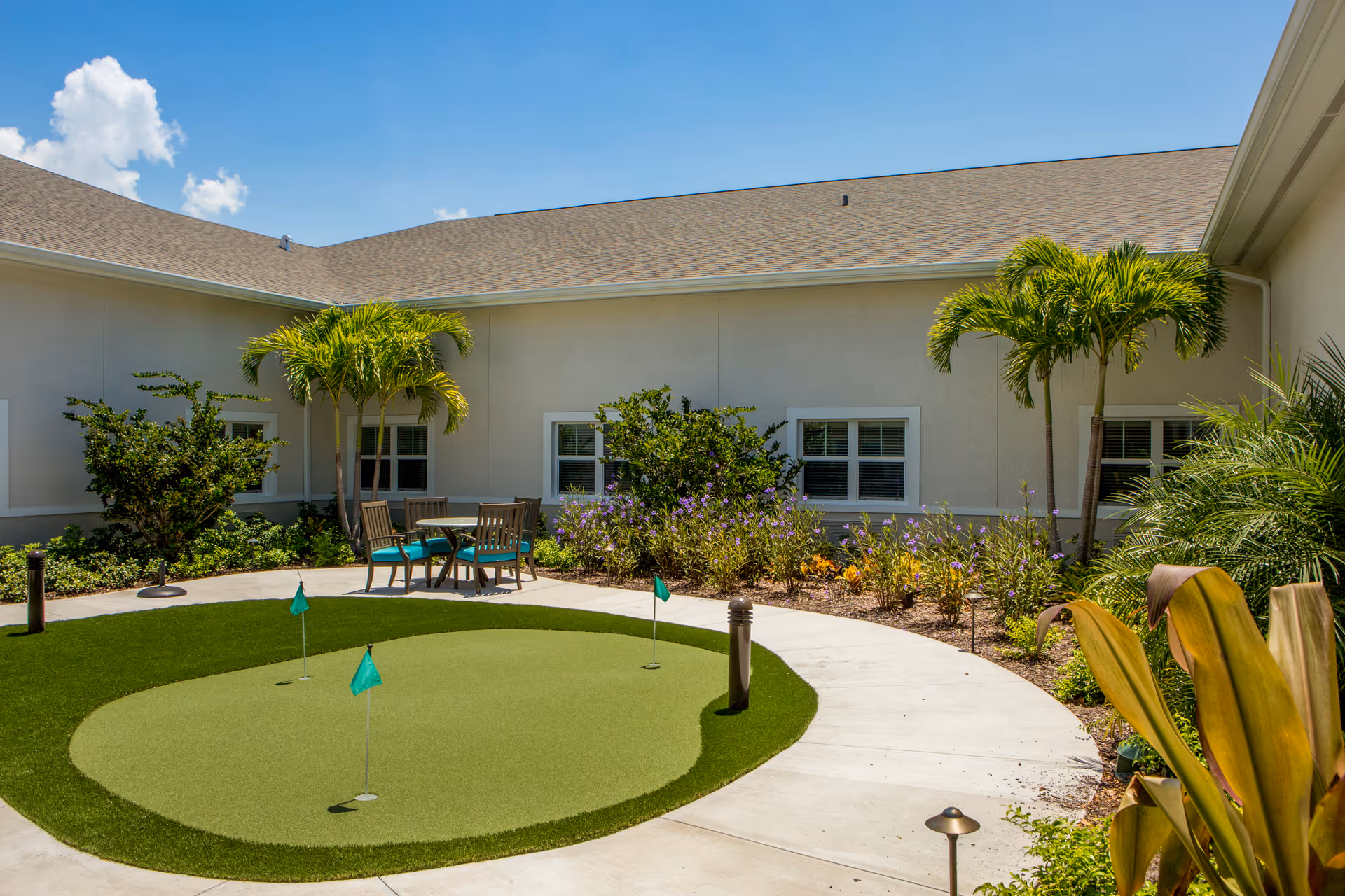 Sunny courtyard with a small putting green, patio table and chairs, palm trees, and landscaped planting beds beside a single-story building.