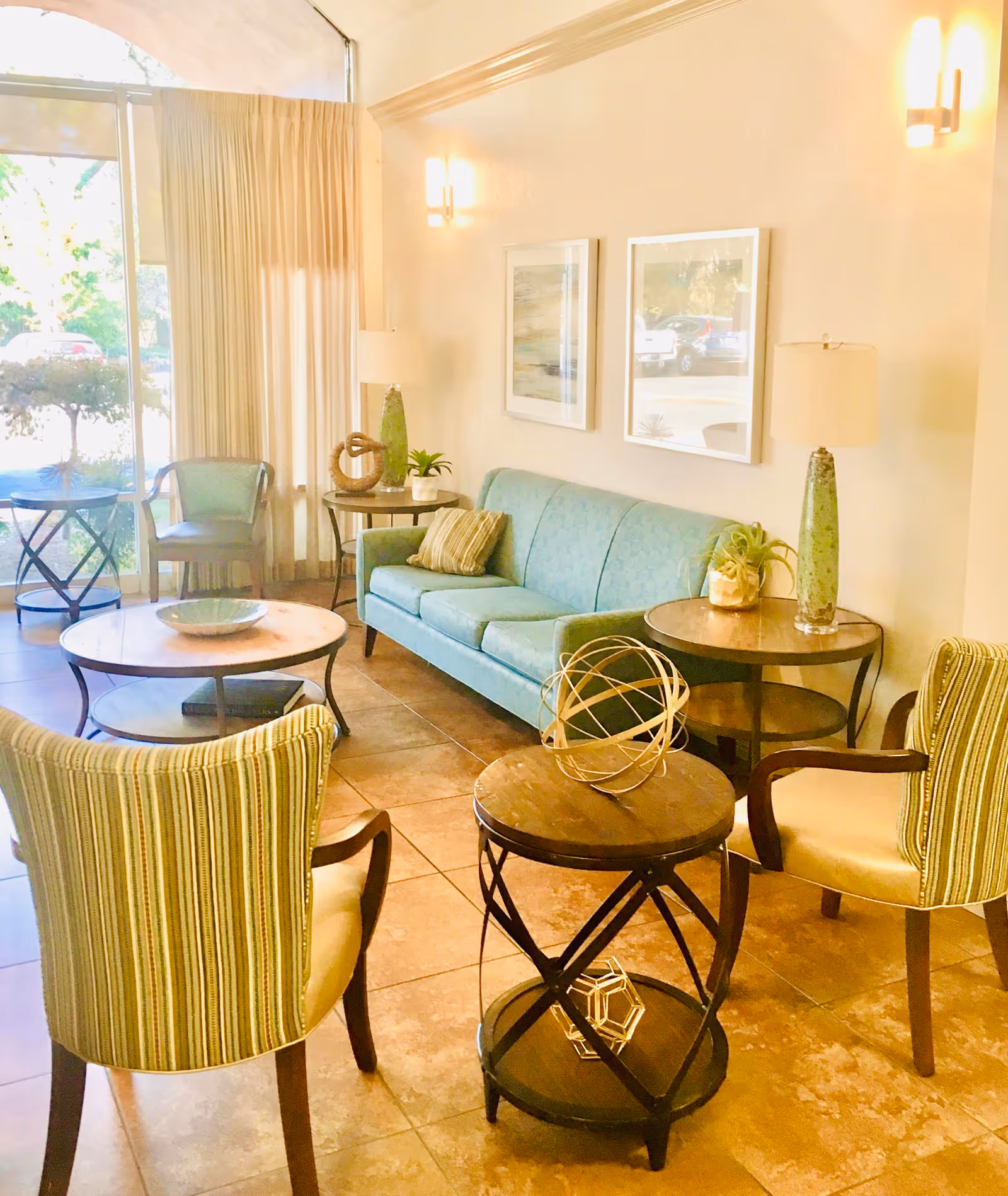 A bright and cozy living room area with a light blue sofa, two striped armchairs, and several round wooden tables. The room has large windows with sheer curtains allowing natural light to fill the space. Decorative items and lamps are placed on the tables and walls.