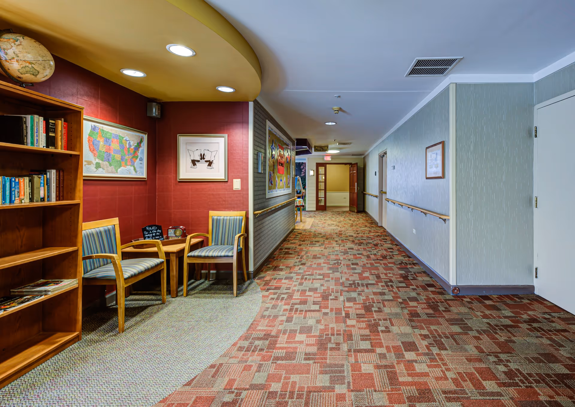 A hallway in a senior living facility with patterned carpet and handrails along the walls. On the left side, there is a small seating area with two striped chairs and a wooden table, a bookshelf filled with books, a globe on top, and framed artwork on the wall. The hallway extends into the distance with doors and bulletin boards visible.