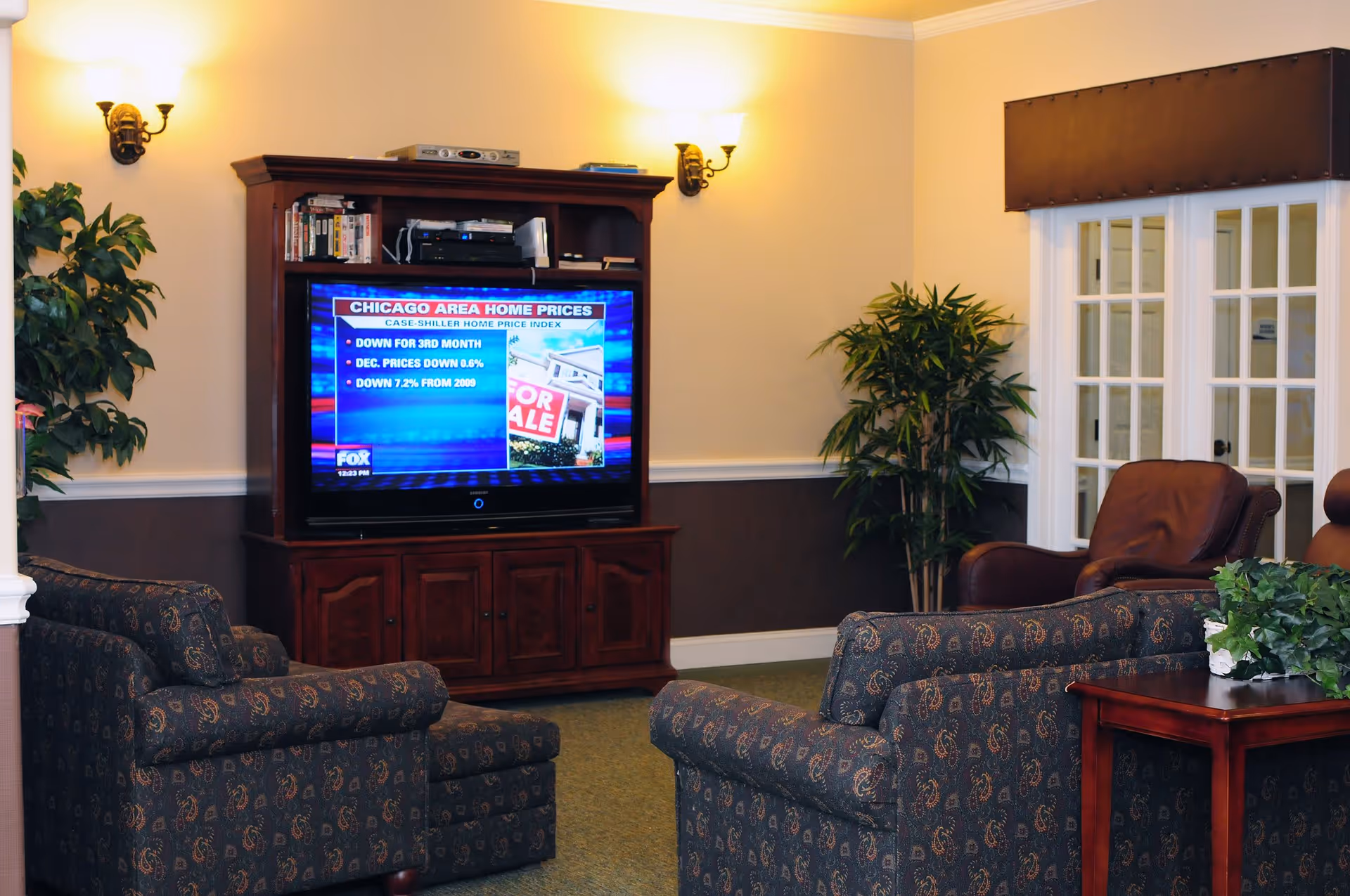 A cozy living room area with patterned armchairs and a leather chair arranged around a wooden entertainment center holding a television. The TV screen displays a news segment about Chicago area home prices. The room is softly lit by wall sconces and decorated with potted plants and a side table with greenery.