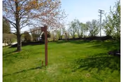A grassy lawn with a small signpost and a leafy tree, bordered by shrubs and utility poles in the background.