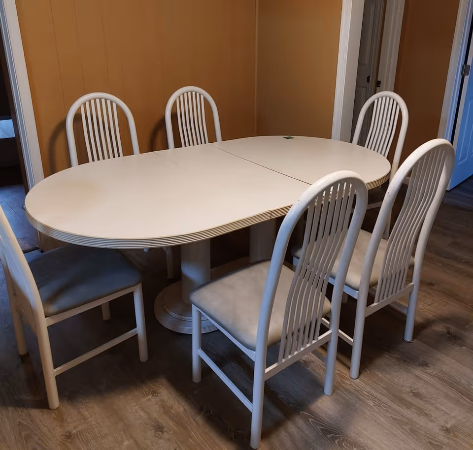 A dining area with a white oval dining table surrounded by six white chairs with vertical slats on the backrests. The room has wood flooring and tan-colored walls. There are doorways visible in the background.