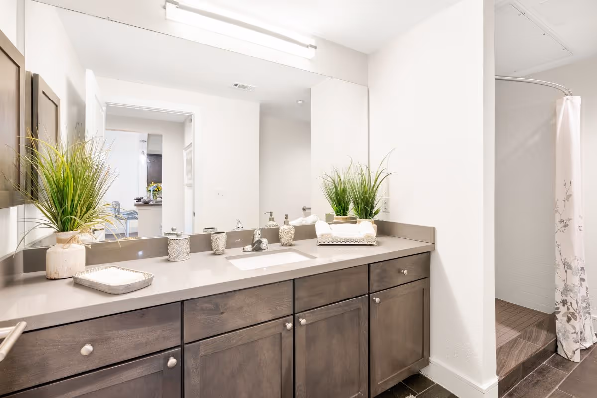 A bright bathroom with a large mirror above a gray countertop and wooden cabinets. The countertop has decorative items including two potted plants, soap dispensers, and a tray with towels. To the right, there is a walk-in shower with a floral-patterned shower curtain. The bathroom has white walls and dark tiled flooring.