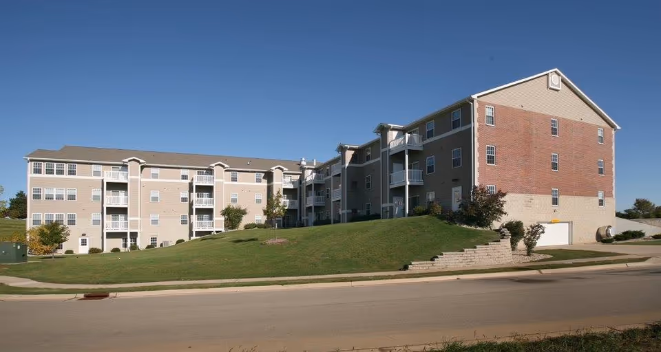 Exterior view of a multi-story senior living facility building with beige and brick siding, balconies, and a well-maintained grassy hill in front under a clear blue sky.